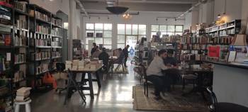 Εσωτερικός χώρος της βιβλιοθήκης, We Need Books, με ανθρώπους που κάθονται γύρω από τραπέζια, διαβάζοντας βιβλία.